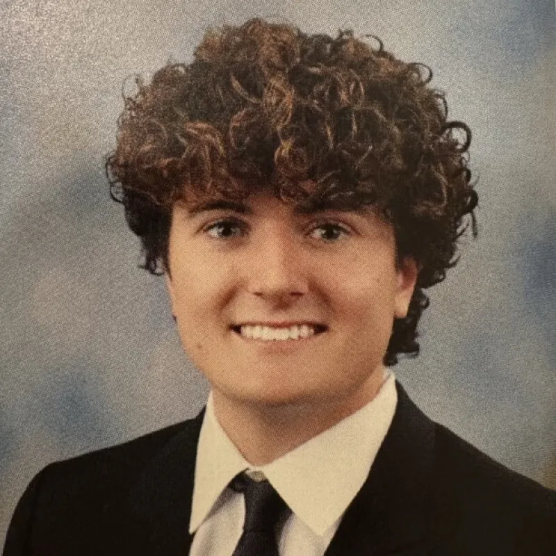 Headshot of a young man with curly brown hair, smiling in a suit and tie.