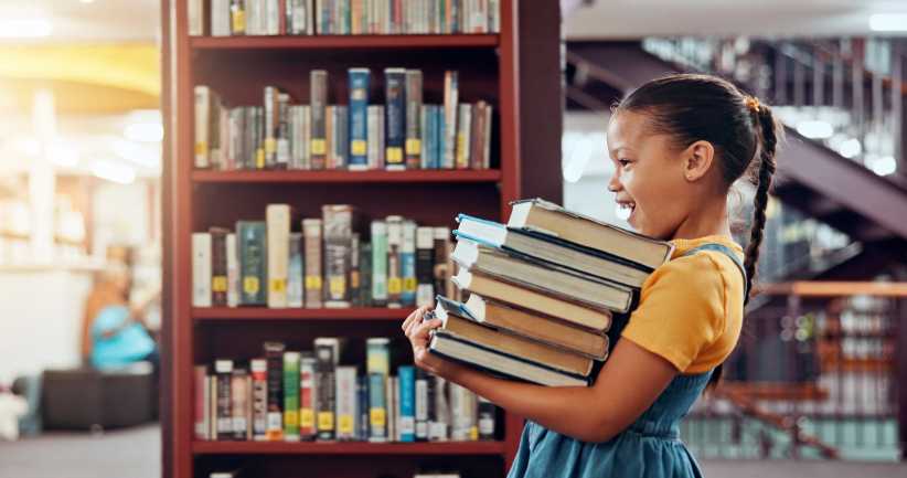 Little girl walking through a library aisle holding a stack of colorful children’s books
