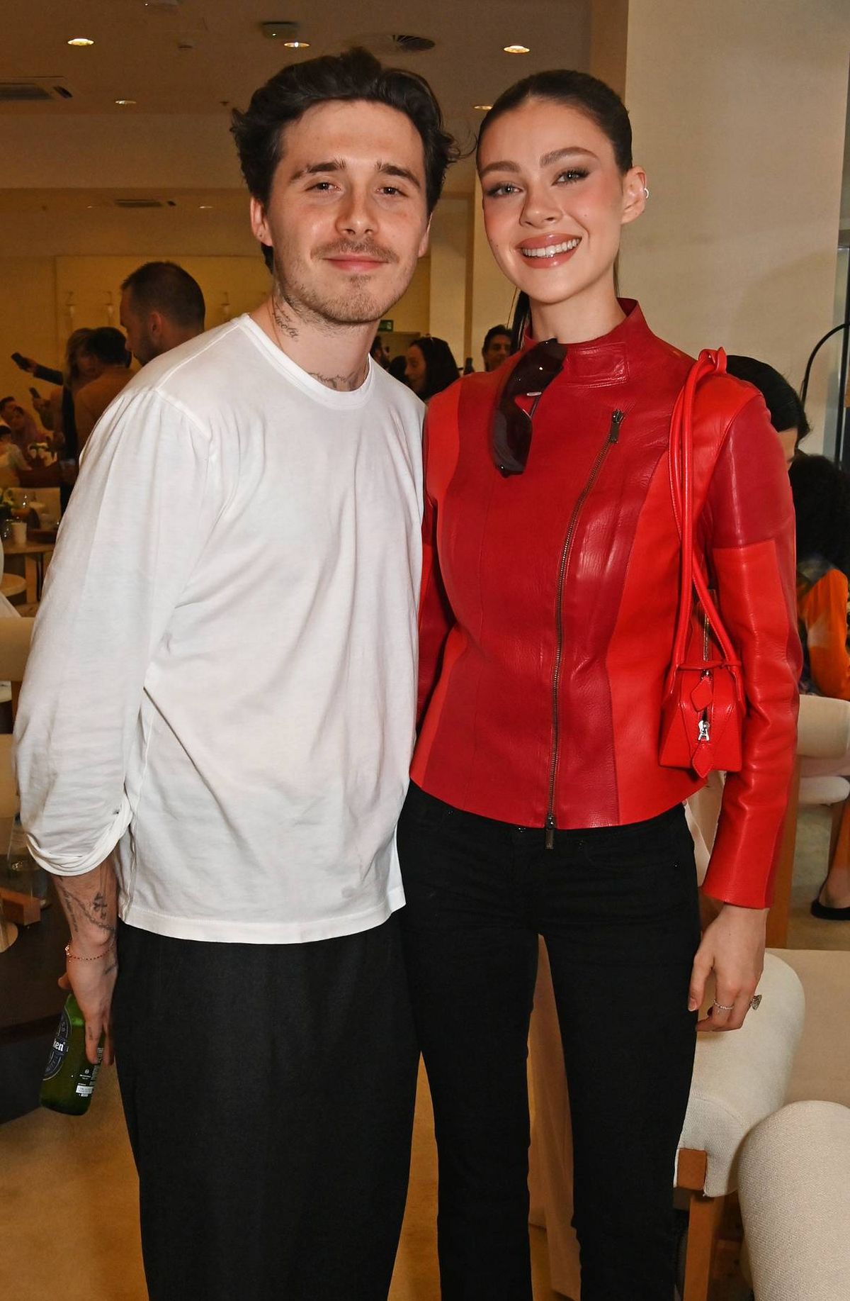 Two individuals, one dressed in a white t-shirt and the other in a red leather outfit, are standing next to each other and smiling for a photograph. The background shows a blurred view of other people and an indoor setting.