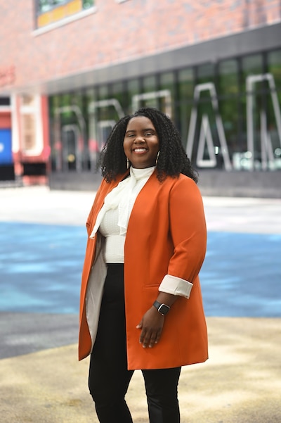 A woman in an orange blazer stands in front of a brick building.