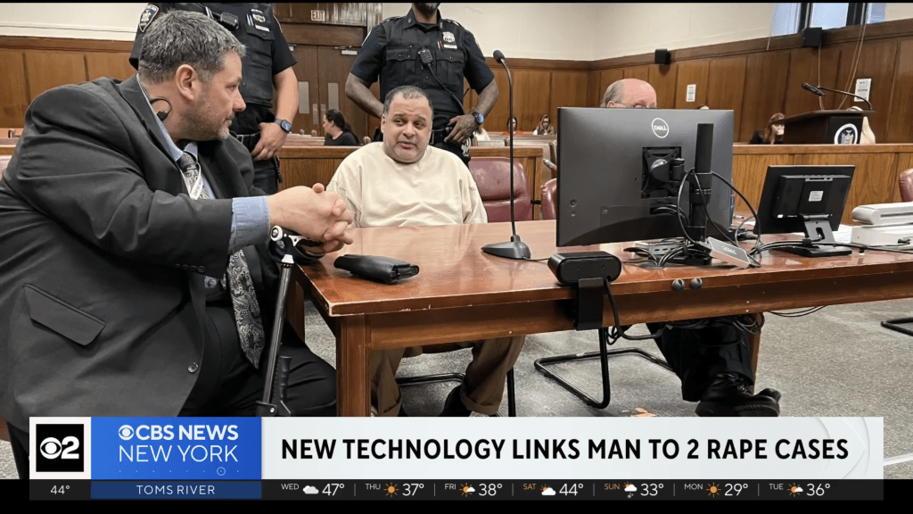 Jancys Santiago, in a light shirt, seated at a court table with his lawyer in a suit and police officers behind them.