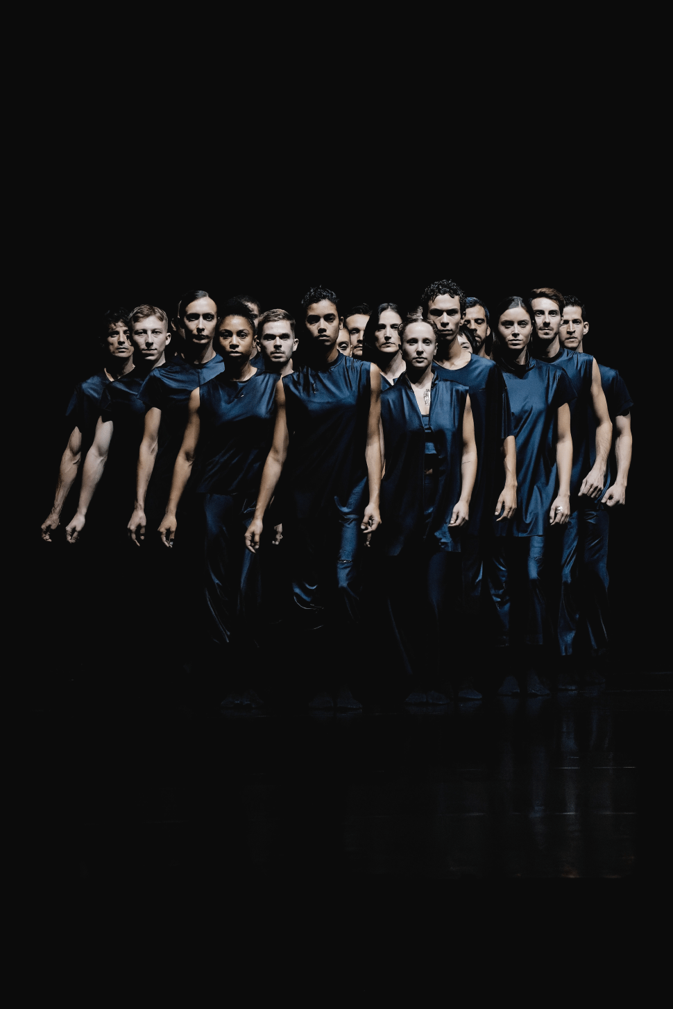 A large ensemble of dancers dressed in dark blue sleeveless costumes stands shoulder to shoulder in a tight formation against a black background, facing forward under dramatic stage lighting.