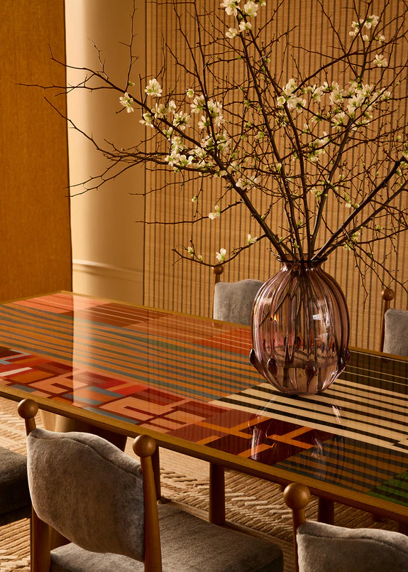 Decorative table with a colorful geometric design, large vase, and blossoming branches, surrounded by gray chairs.