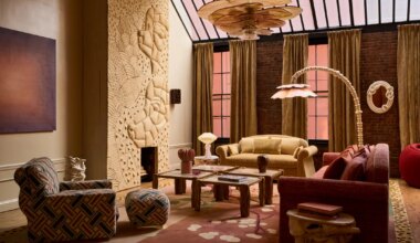 Eclectic living room with patterned furniture, carved wall art, large chandelier, and tall windows with brown curtains.