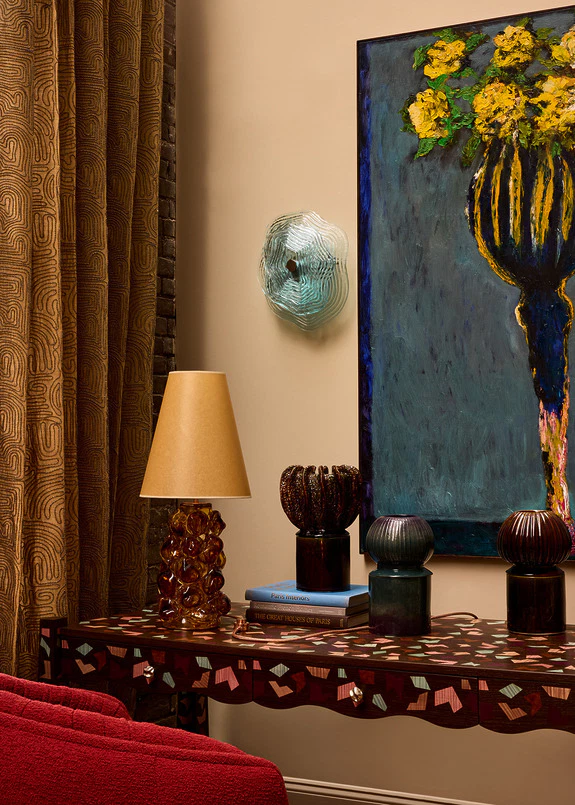Eclectic living room corner with a decorative lamp, abstract painting, books, and ornate table against a textured curtain backdrop.
