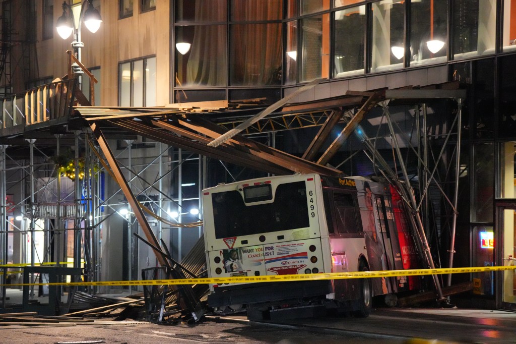 The rear of a Coach USA bus is seen partially under collapsed building scaffolding, with yellow caution tape in the foreground.