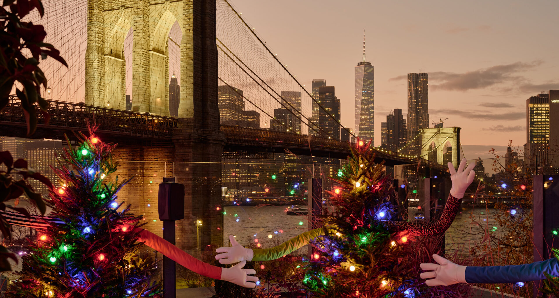 A rooftop installation of Christmas trees at DUMBO House, each tree decorated with multicolored lights and playful, fabric-covered arms reaching outward, set against sweeping views of the Brooklyn Bridge and the Manhattan skyline at dusk.