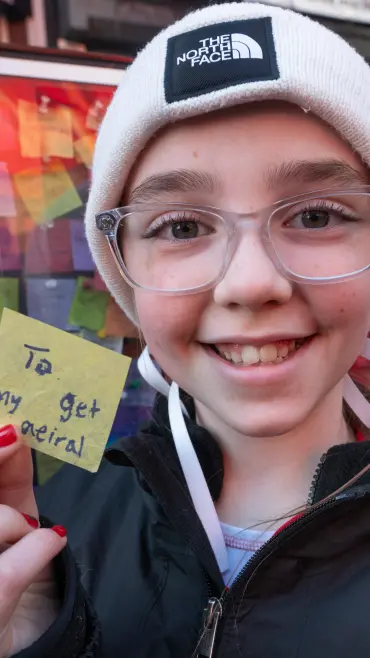 Leah Frisbie holding a yellow sticky note that reads "To my get aerial" in front of the Times Square Wishing Wall.