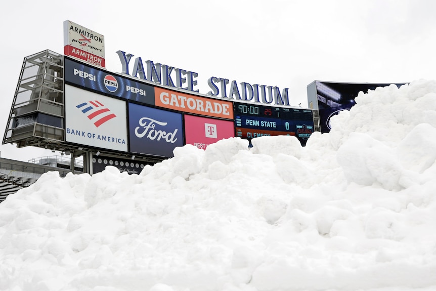A large pile of white snow is piled up on the ground. In the background is a billboard that reads 'Yankee Stadium'. 