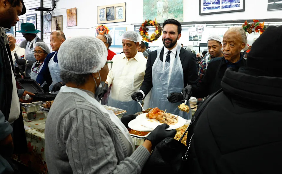 The Rev. Al Sharpton, right, and New York Mayor-elect Zohran Mamdani, in a suit underneath an apron, serve Thanksgiving meals in Harlem on Nov. 27, 2025.