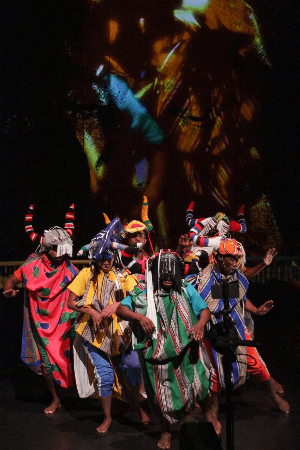 A group of dancers wearing brightly colored, patterned costumes and horned headpieces perform barefoot on a dark stage with an abstract, multicolored projection behind them.