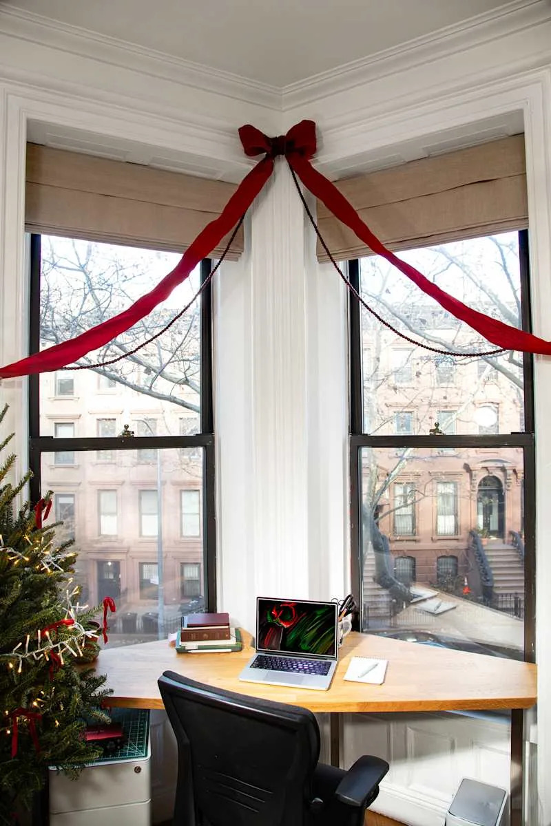 Cozy home office corner with a wooden desk, laptop, books, and a decorated Christmas tree. Red ribbons adorn the windows.