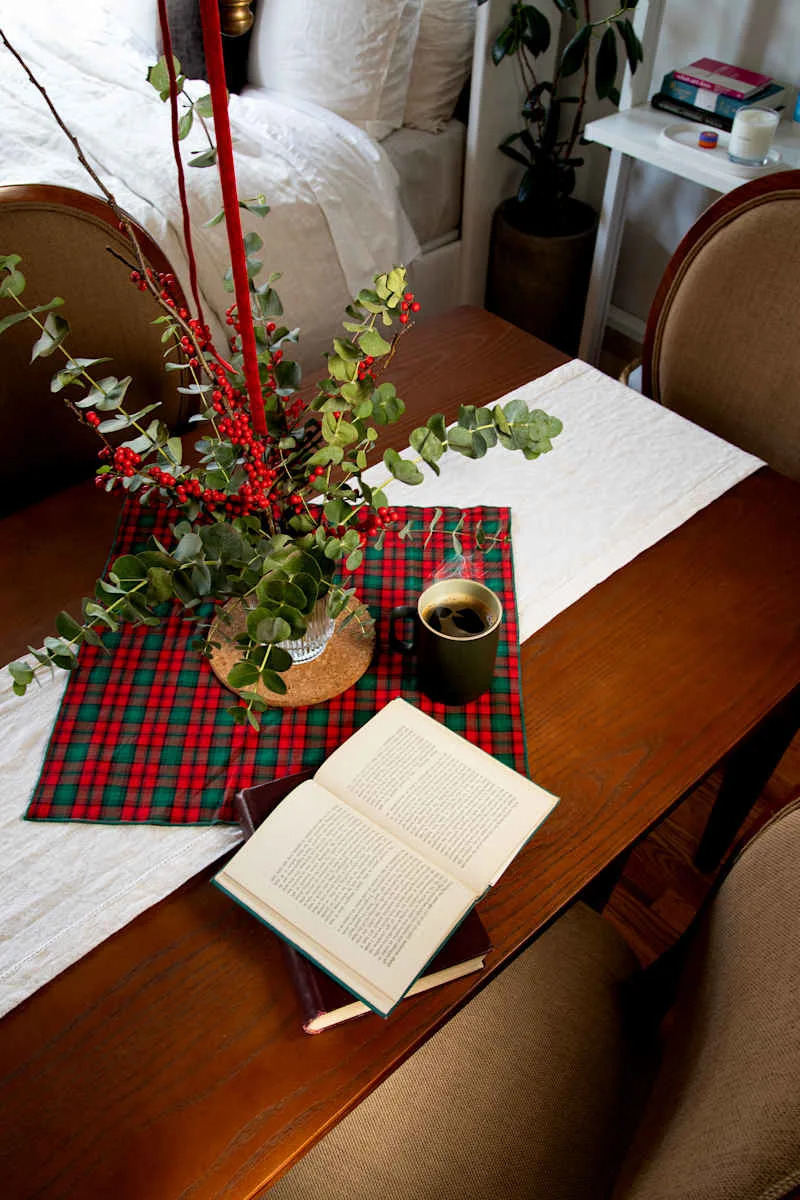 A wooden dining table set with a plaid tablecloth, a floral arrangement, an open book, and a coffee mug.