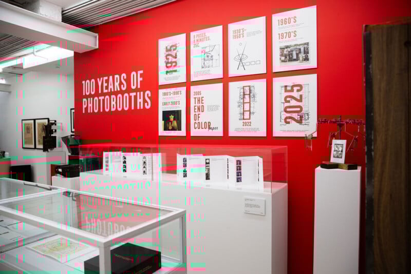 Museum exhibit with a bright red wall displays "100 Years of Photobooths" and a timeline with dates and images. Glass cases showcase photostrips, historical artifacts, and vintage photo equipment.