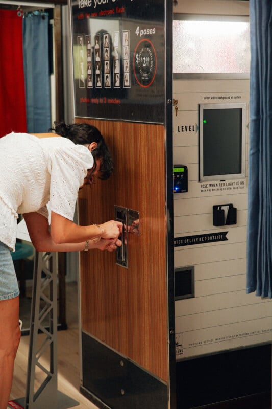 A woman in a white blouse and denim shorts bends down to insert coins into a vintage photo booth with wood paneling, preparing to take photos.