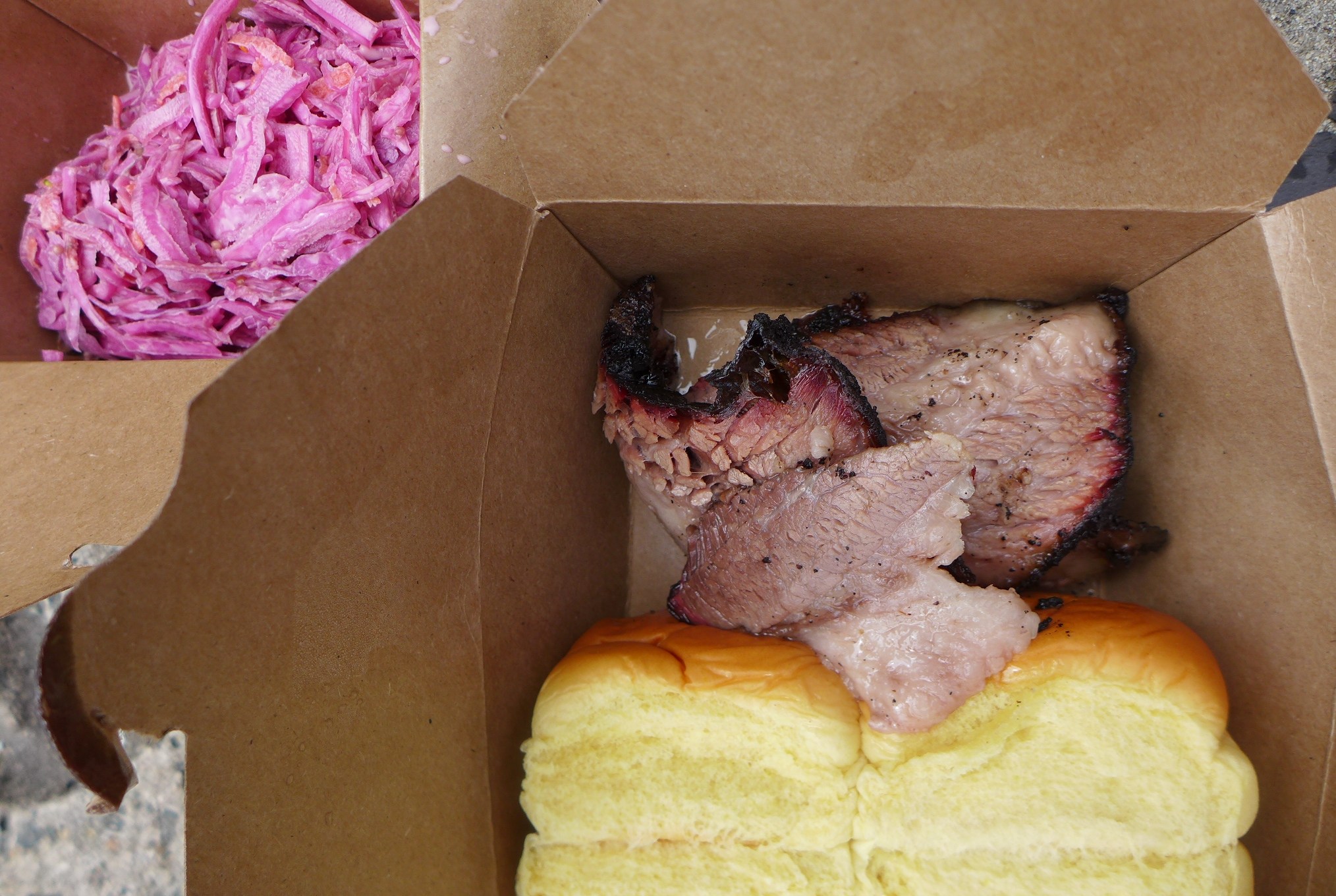 A brown cardboard box with sliced brisket and rolls seen from above, with pink cole slaw in the side.