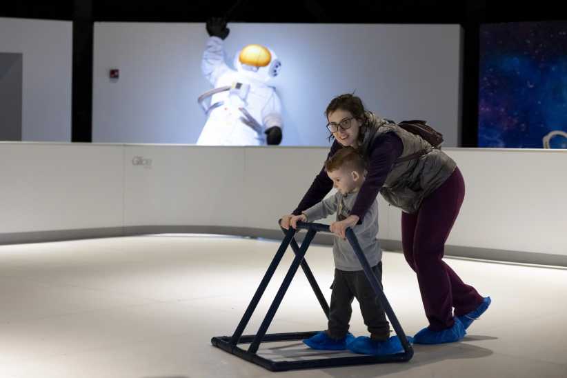 A young mom leaning over her child as they hold onto a balancing object while they skate in the new skating experience Space Glide at New York Hall of Science