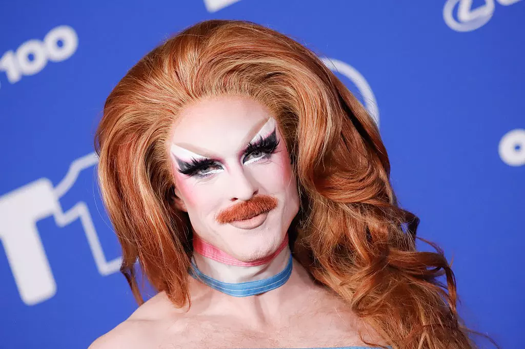 A performer in a sparkly orange, white, and black outfit with a flowing cape poses dramatically on stage wearing white heeled boots.