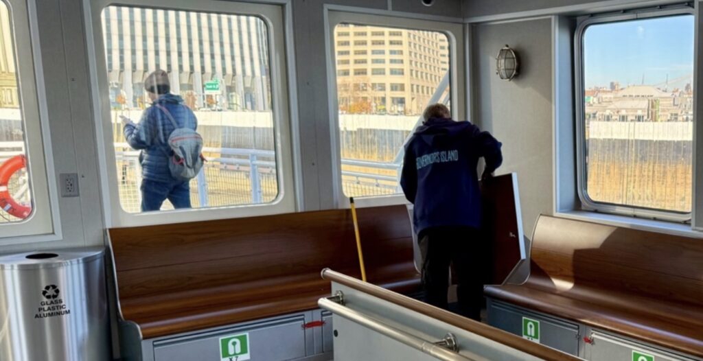 Hamilton, crew member, prepares the cabin of the Governors Island ferry between runs as a passenger walks along the dock outside. (Credit: Baron Carr)