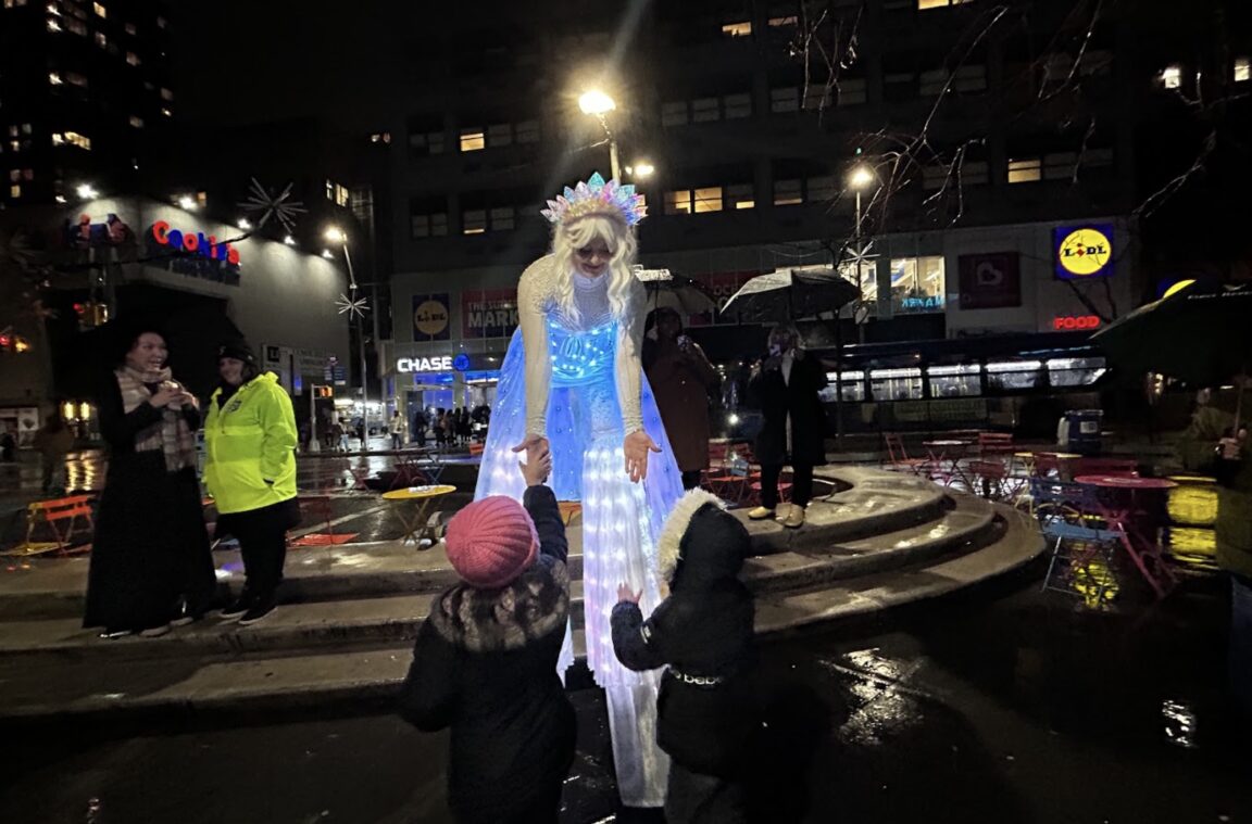Brianna Kalisch, a circus variety performer, greets two children during a tree lighting ceremony on a rainy night in Downtown Brooklyn. (Credit: Loyalty Jean Aimé)