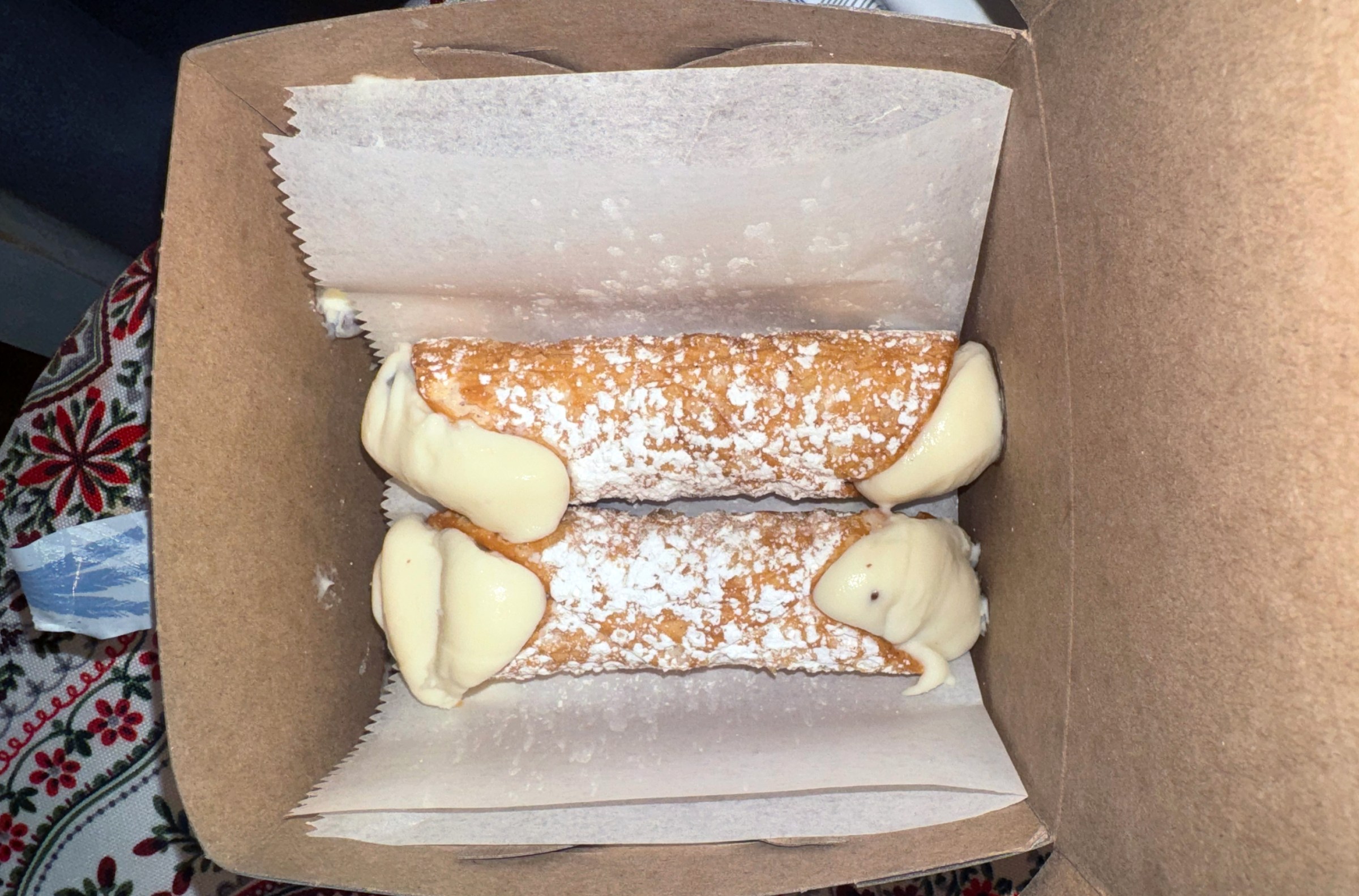 Two cannoli on wax paper in a brown cardboard box on someone’s lap.