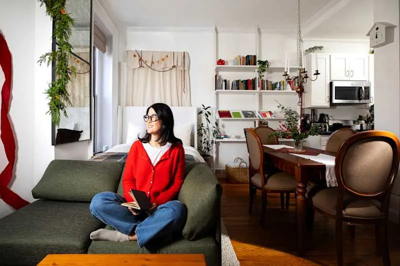 A woman in a red cardigan sits on a green sofa, holding a notebook, in a cozy living space with a dining area and bookshelves.