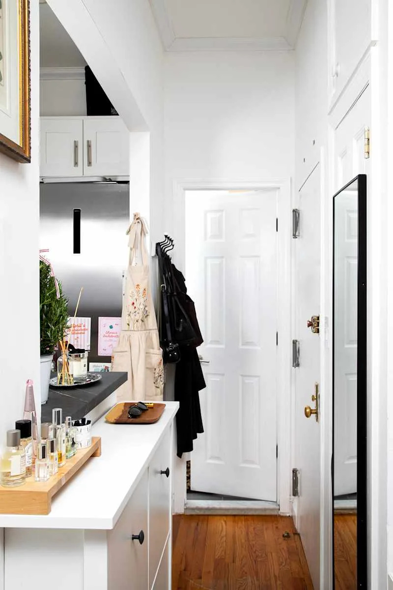 Narrow entryway with white walls, a mirror, a black countertop, and a wooden tray holding perfumes and sunglasses.