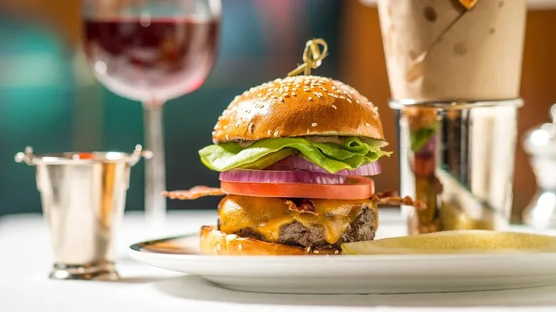 A burger at The Polo Bar sitting on a plate with a pickle and a cup of french fries wrapped in paper.