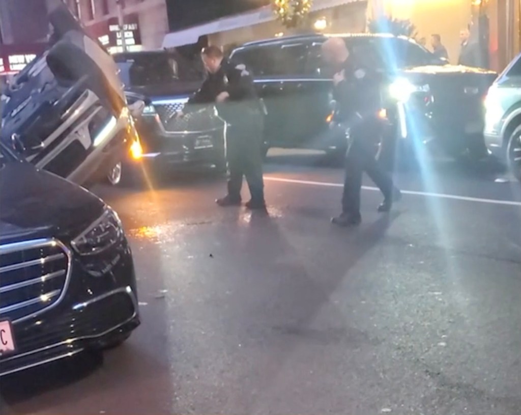 A black car, possibly a Porsche, is overturned on a city street, with two police officers standing nearby.