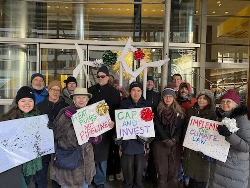 A dozen NY Sierra Club members hold holiday signs and paper windmills outside Governor Hochul's office.