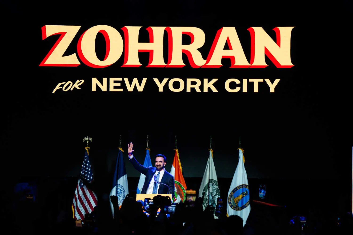 Zohran Mamdani raises his right hand as he smiles beneath a giant “Zohran for New York City” sign on election night.