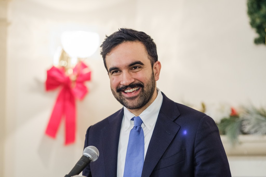 New York City Mayor-elect Zohran Mamdani smiling while standing at a microphone.