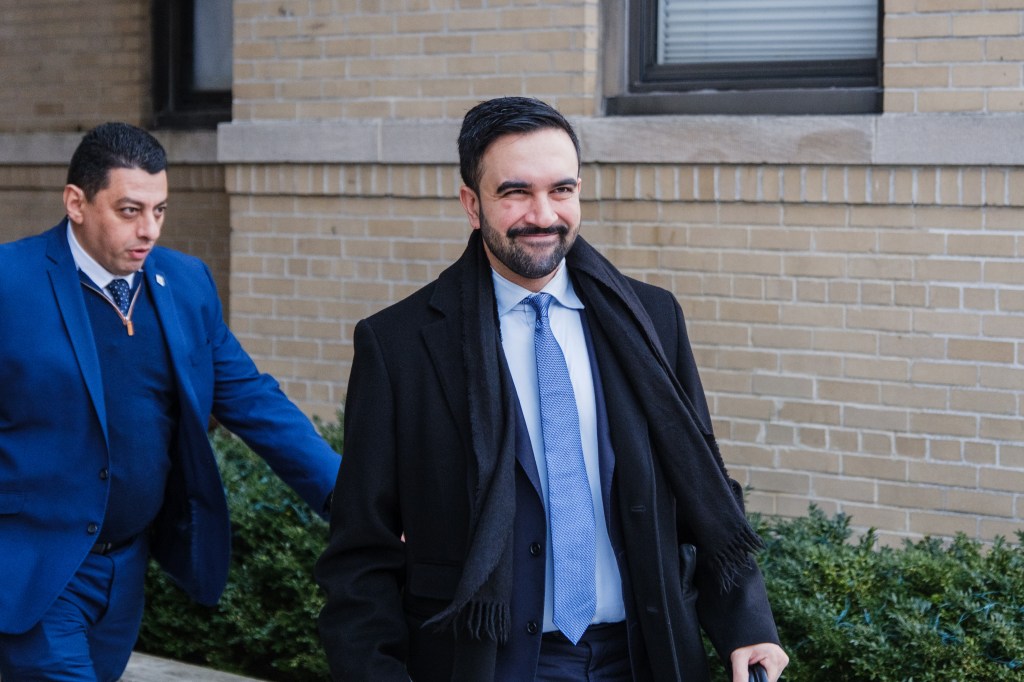New York City mayor-elect Zohran Mamdani smiles while leaving his apartment in Astoria, Queens, accompanied by another man in a blue suit.