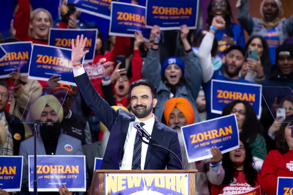 Zohran Mamdani waves to a cheering crowd holding "Zohran for New York City" signs at a rally.