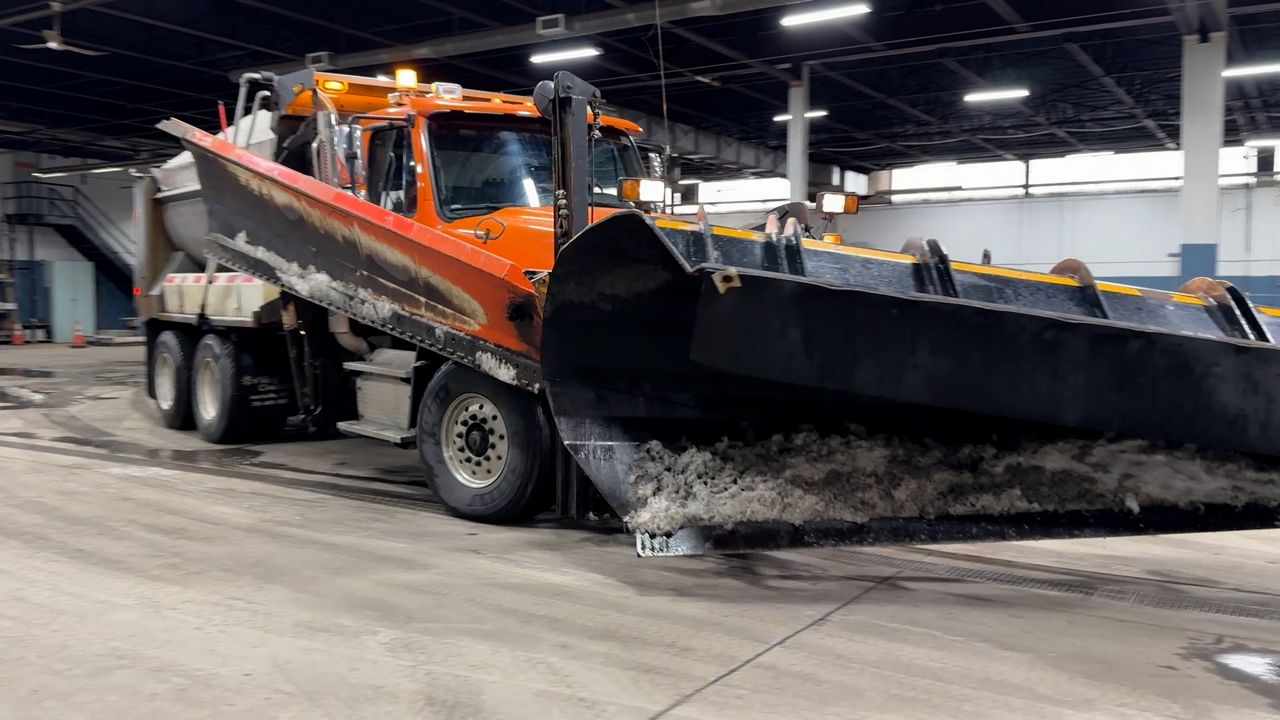 Orange and black snow plow truck with large front blade attachment parked inside a maintenance facility garage.
