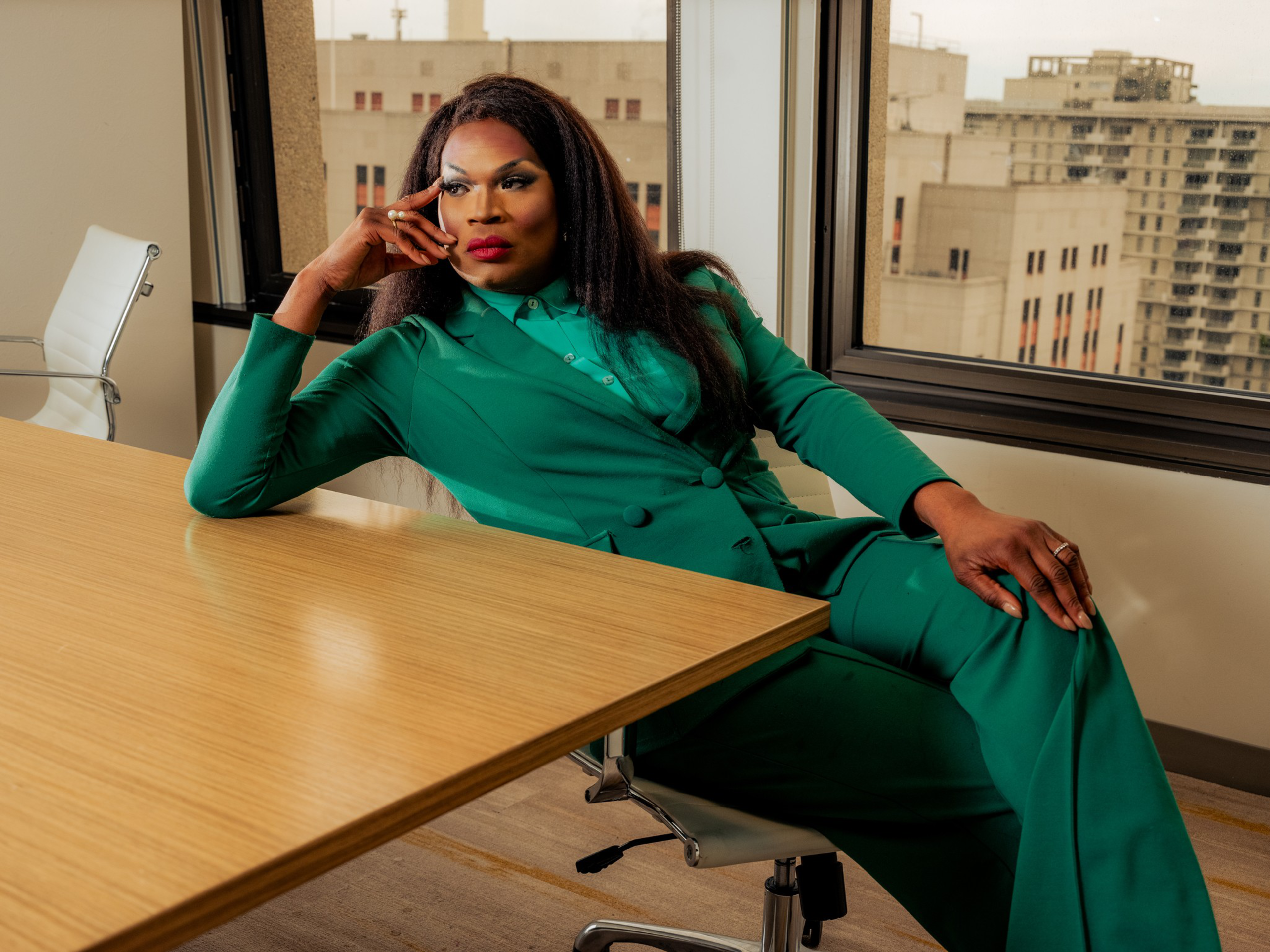 A person in a green suit reclines confidently in an office chair, resting their head on one hand with city buildings visible through the windows behind them.