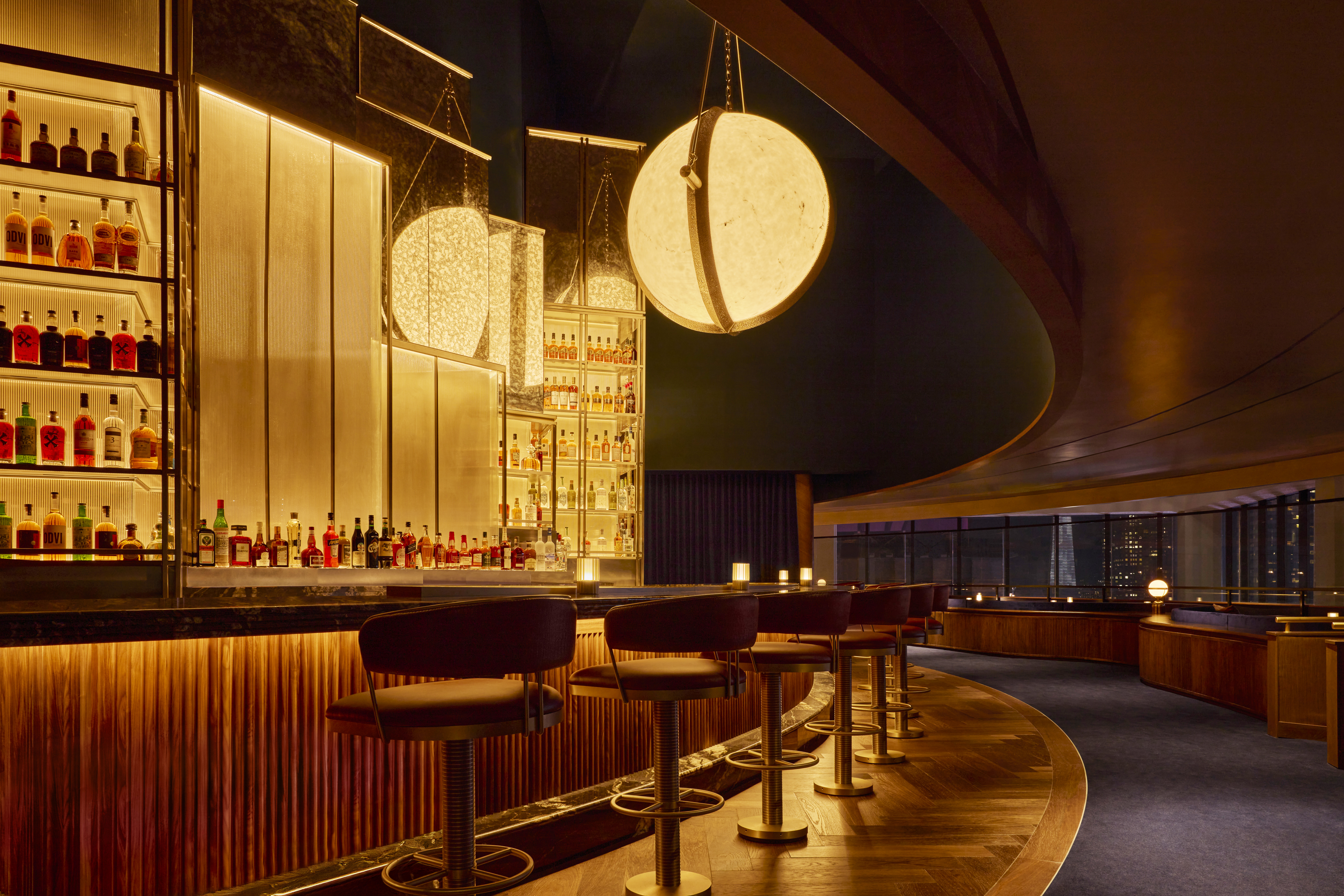 A dimly lit bar with a long, curving counter lined with red-brown stools, a tall liquor display, and an ornate overhead light fixture.