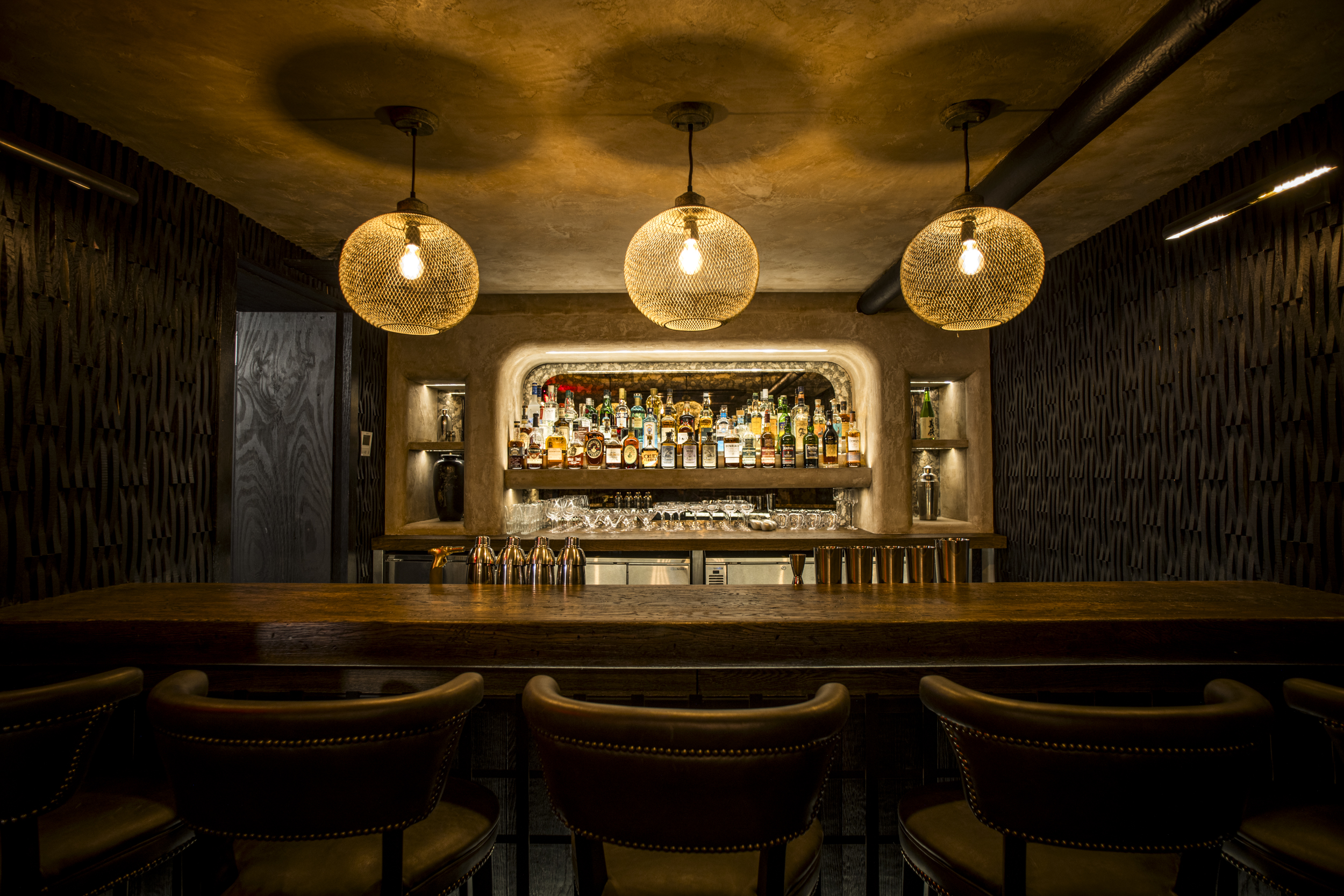 The interior of the Sip & Guzzle cocktail bar, featuring a wooden bar, barstools, and a backlit shelving unit with various liquor bottles.