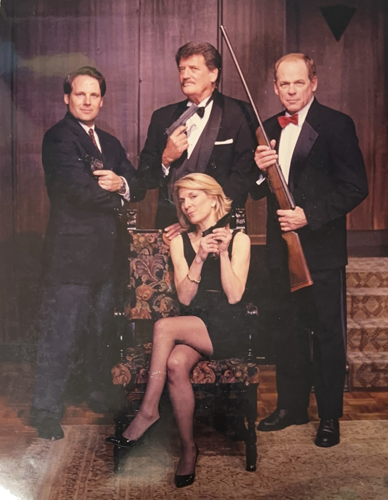 Cast photo for the off-Broadway play "Perfect Crime," with three men in suits holding guns and a woman in a black dress seated in a patterned chair, holding a pistol.