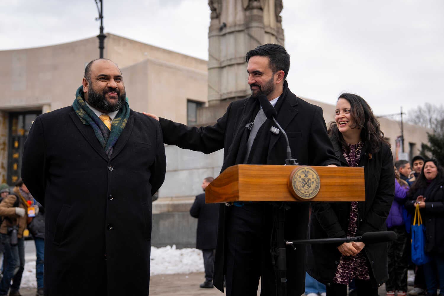 Mayor Zohran Mamdani, wearing a dark overcoat and standing behind a lectern, puts his hand on the shoulder of Ali Najmi, his choice to chair the Mayor’s Advisory Committee on the Judiciary. Mr. Najmi is wearing a dark coat and bright yellow tie. They are standing at Grand Army Plaza in Brooklyn.
