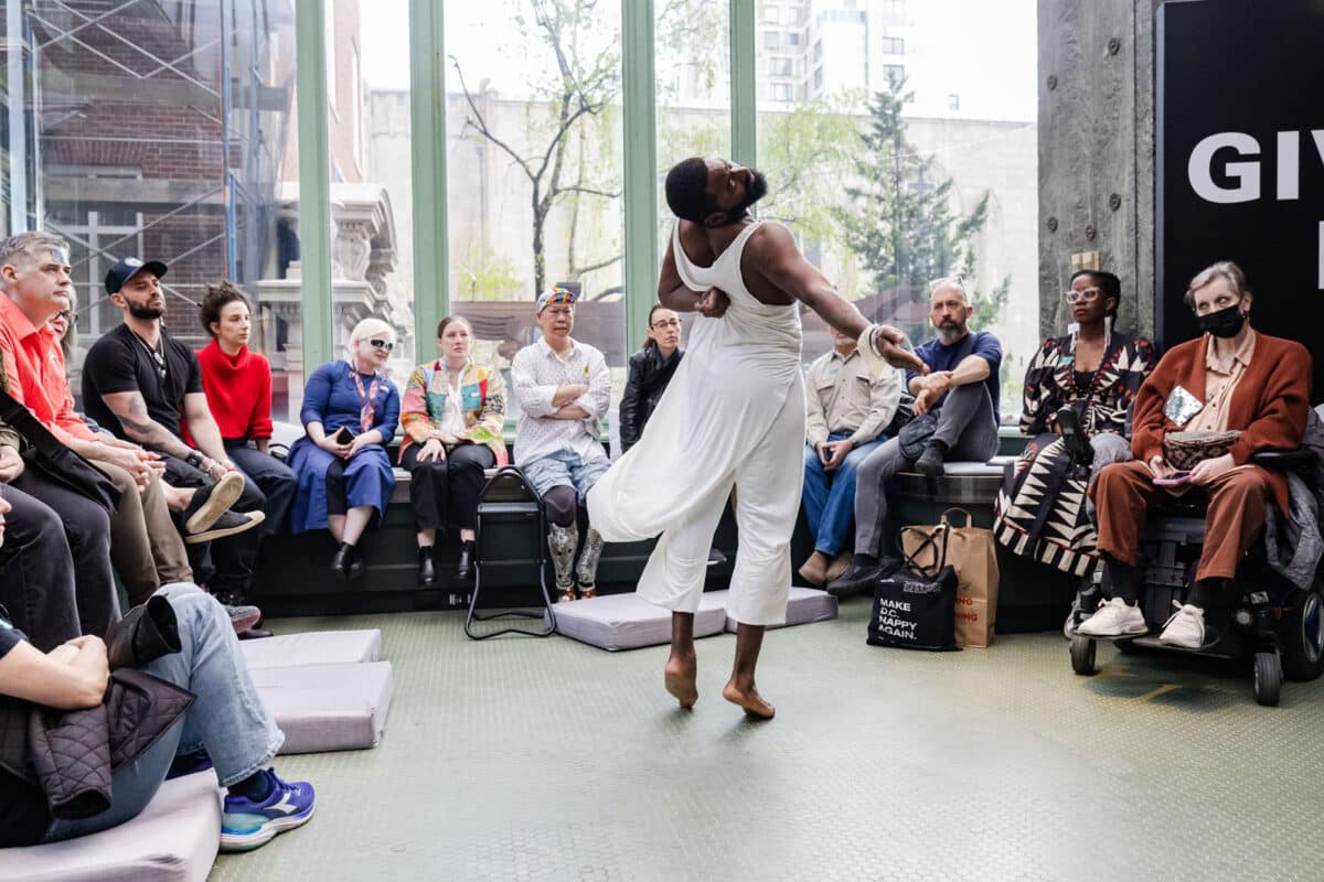 Alameda Post - Jerron Herman twirls in mid-turn dressed in a long, flowy white tunic in front of the audience in a light-filled atrium. 