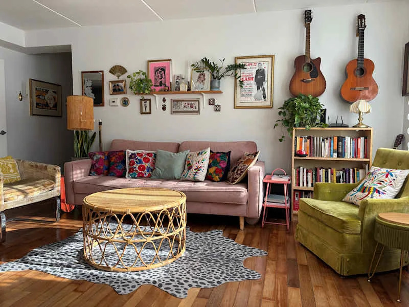 Cozy living room with a pink sofa, colorful cushions, a rattan coffee table, green armchair, and bookshelves.