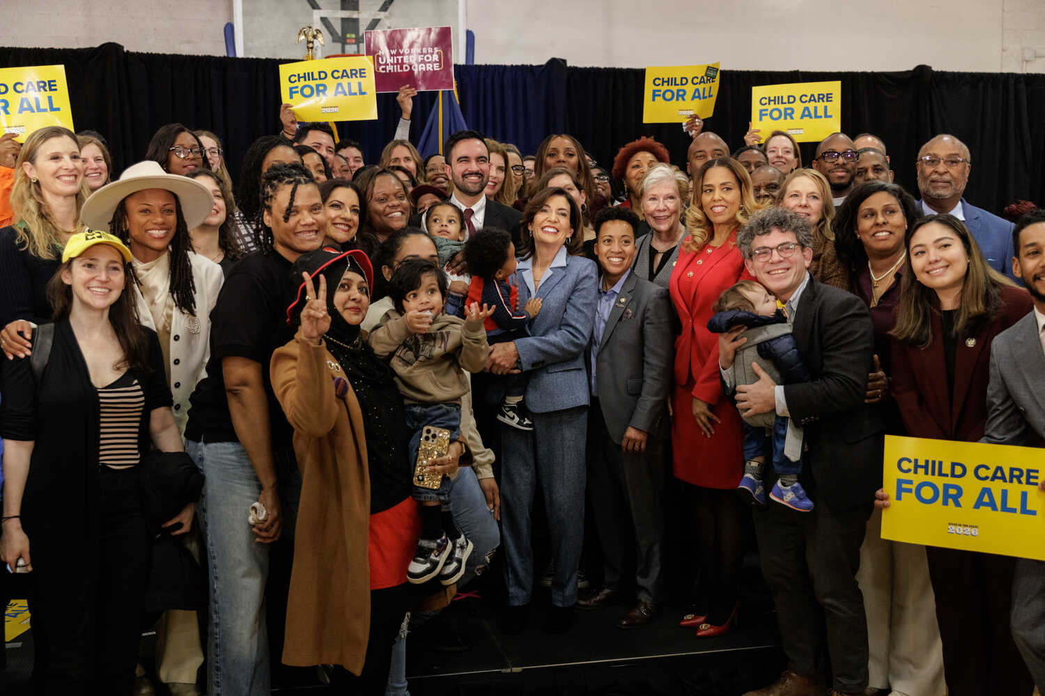 Lawmakers and advocates, some holding children and others holding signs that say Child Care For All, stand around Gov. Kathy Hochul and Mayor Zohran Mamdani.