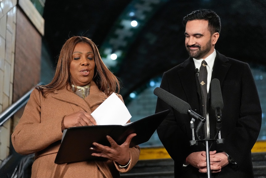 Letitia James flicks through a binder of notes as Zohran Mamdani stands next to her and smiles
