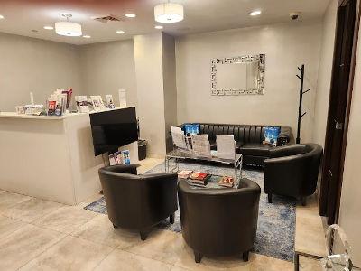 This professional reception space features a long black leather tufted sofa accented with blue decorative pillows and three matching black leather tub chairs. The seating is arranged around a glass-topped coffee table with a silver frame, set atop a blue