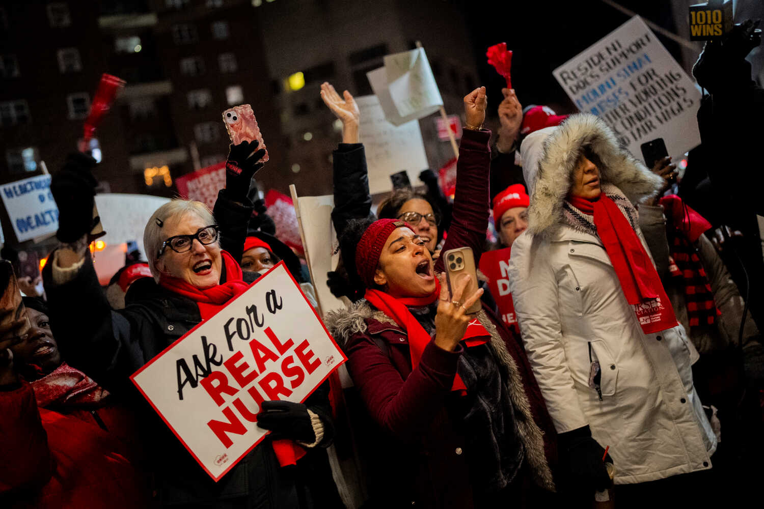 A group of women dressed in winter coats wearing red scarves chant and hold signs, one reading "Ask for a REAL NURSE"