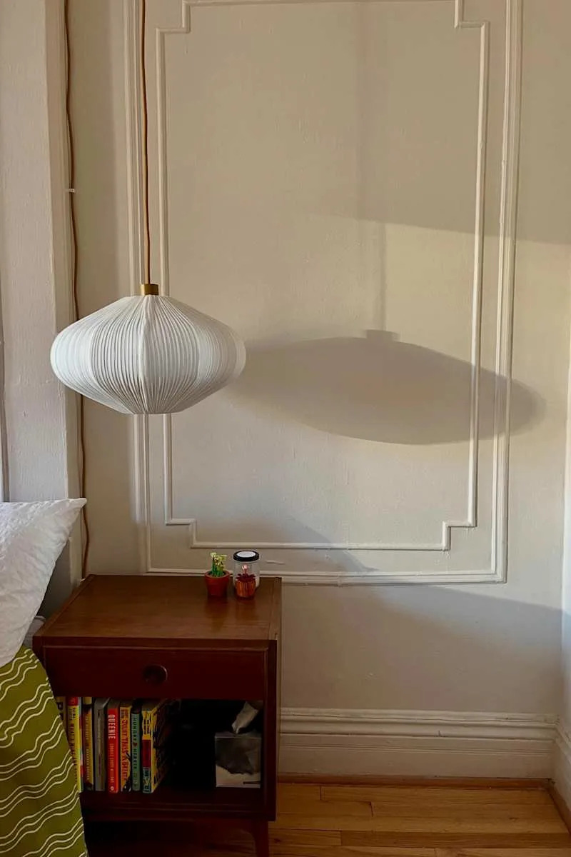 A white pendant lamp casts a shadow on a wall above a wooden nightstand with books, small plants, and a jar.