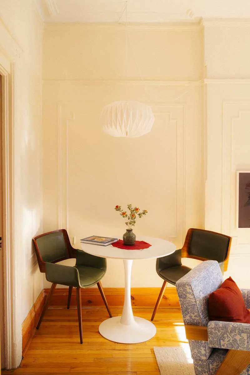 Cozy dining nook with a round white table, two green chairs, a vase of flowers, and a patterned blue sofa.