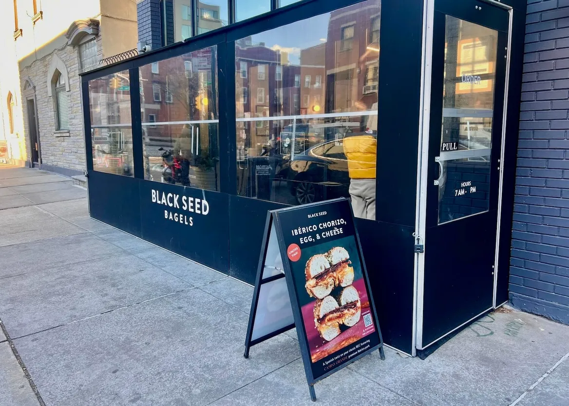 Black storefront with three large windows, a door, and a sidewalk sign advertising bagels