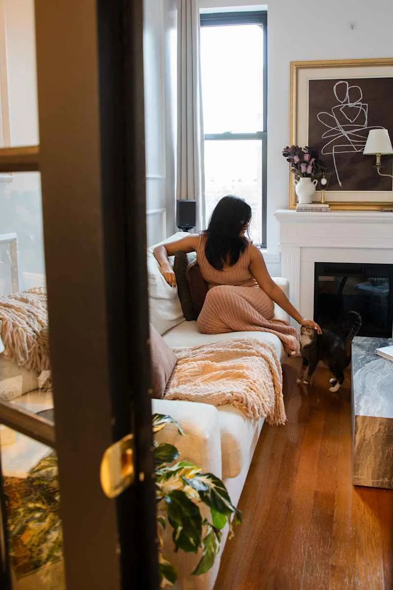 A woman in a pink dress sits on a light sofa, petting a cat, with a cozy throw and a marble coffee table nearby.